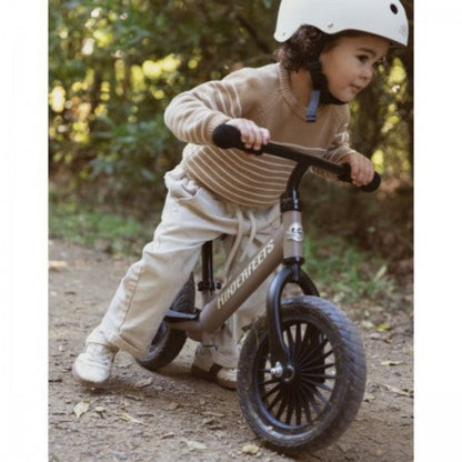 Young child wearing a white helmet riding a beige Kinderfeets balance bike with black wheels and handlebars along an outdoor dirt path.